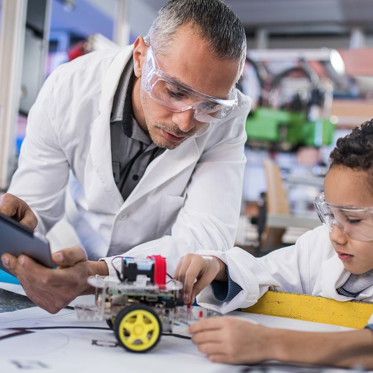 Mid adult teacher using touchpad while looking at small boy making a robot in a laboratory. Focus is on man.